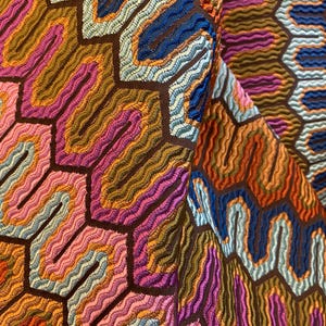 May include: Close-up of a colorful fabric with a geometric pattern. The design features a series of interconnected, hexagonal shapes filled with wavy lines in shades of orange, pink, blue, and brown. The fabric has a textured appearance.