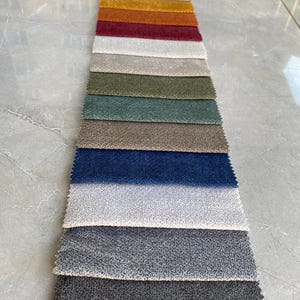 Op de afbeelding: Een stoffen stalenkaart met een verscheidenheid aan gestructureerde bekledingsstalen. De stalen tonen een reeks kleuren, waaronder neutrale tinten, warme oranjetinten, diepe roden en koele blauwen. De stof lijkt een zacht materiaal te zijn, geschikt voor meubels.
