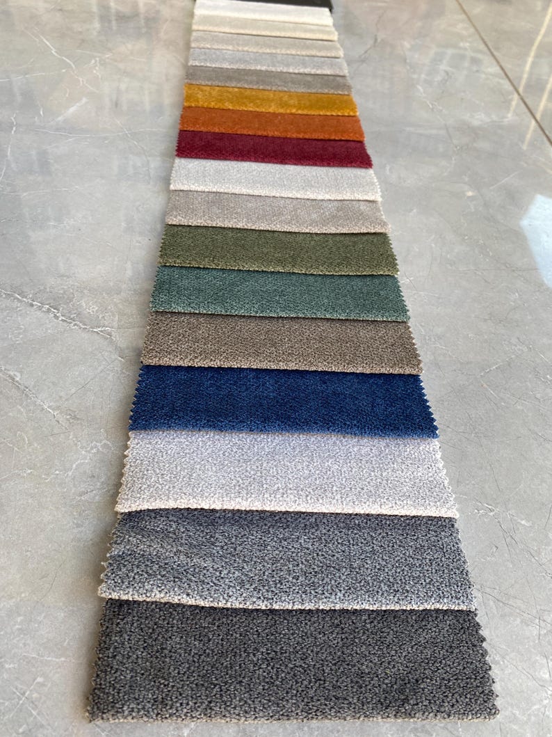 Op de afbeelding: Een stoffen stalenkaart met een verscheidenheid aan gestructureerde bekledingsstalen. De stalen tonen een reeks kleuren, waaronder neutrale tinten, warme oranjetinten, diepe roden en koele blauwen. De stof lijkt een zacht materiaal te zijn, geschikt voor meubels.