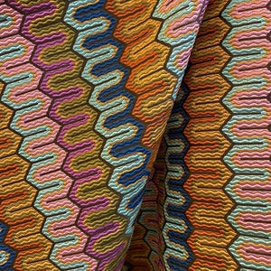 May include: Close-up of a colorful fabric with a geometric pattern. The design features a repeating honeycomb structure with wavy lines in shades of orange, blue, pink, and purple, set against a dark brown background. The fabric is folded, revealing its texture.
