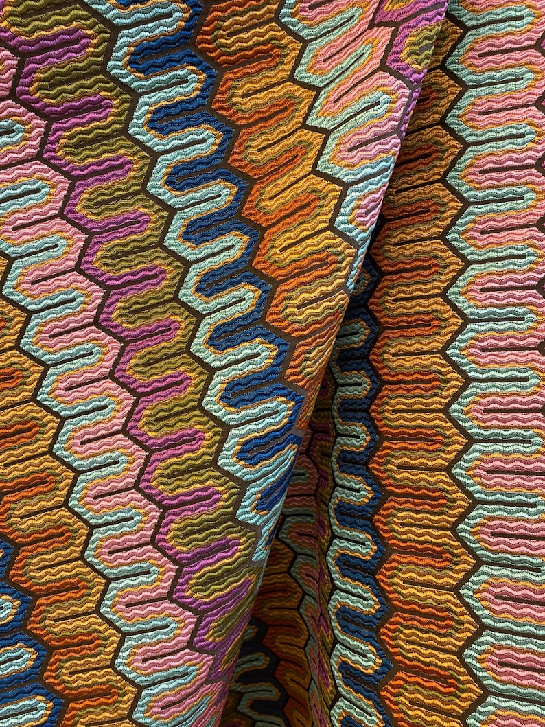 May include: Close-up of a colorful fabric with a geometric pattern. The design features a repeating honeycomb structure with wavy lines in shades of orange, blue, pink, and purple, set against a dark brown background. The fabric is folded, revealing its texture.