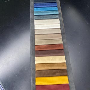 May include: A fabric swatch card featuring a variety of colors. The samples range from light gray to deep teal, with additional shades of beige, brown, and yellow. The card is displayed on a dark surface, showcasing the diverse color palette.