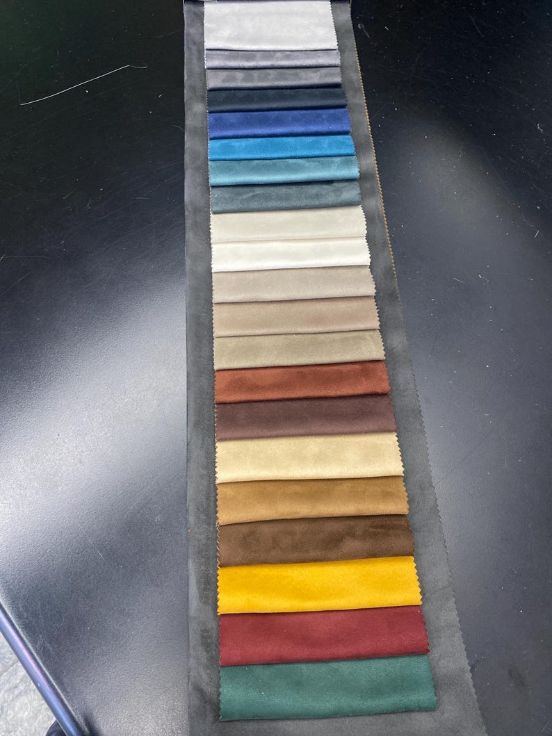 May include: A fabric swatch card featuring a variety of colors. The samples range from light gray to deep teal, with additional shades of beige, brown, and yellow. The card is displayed on a dark surface, showcasing the diverse color palette.