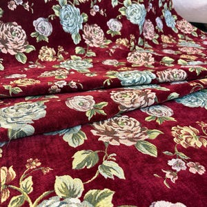 Könnte beinhalten: Burgunderfarbener Stoff mit floralem Muster. Der Stoff zeigt große Rosen in Rosa-, Blau- und Cremetönen mit grünen Blättern. Die Rosen sind über den Stoff verteilt und erzeugen eine Vintage- und romantische Ästhetik.