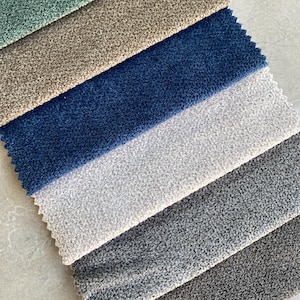 Op de afbeelding: Stalen van stoffen in verschillende kleuren, waaronder teal, beige, marineblauw, lichtgrijs, middengrijs en donkergrijs. De stof heeft een getextureerde, geluste pool, geschikt voor bekleding of interieurprojecten.