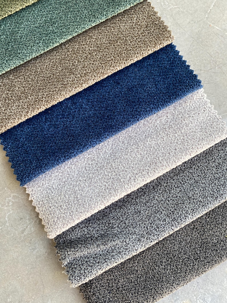 Op de afbeelding: Stalen van stoffen in verschillende kleuren, waaronder teal, beige, marineblauw, lichtgrijs, middengrijs en donkergrijs. De stof heeft een getextureerde, geluste pool, geschikt voor bekleding of interieurprojecten.