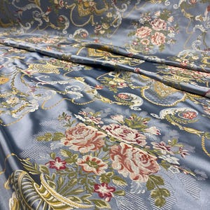 Könnte beinhalten: Nahaufnahme eines gefalteten, seidigen Stoffes in einem gedämpften Blauton. Der Stoff ist mit einem detaillierten Blumenmuster mit Rosen in Rosa-, Creme- und Rottönen sowie goldenen und grünen Akzenten verziert. Das Design hat eine Vintage- und verzierte Ästhetik.
