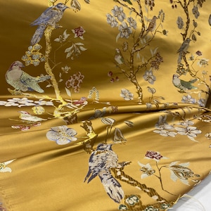 May include: A golden yellow fabric featuring a woven pattern of birds perched on branches with white and blue flowers. The design includes various birds in shades of blue, green, and brown, interspersed with floral motifs across the surface.