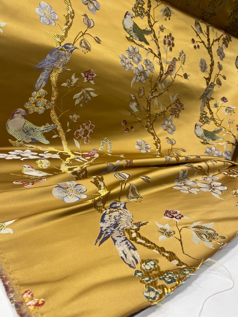 May include: A golden yellow fabric featuring a woven pattern of birds perched on branches with white and blue flowers. The design includes various birds in shades of blue, green, and brown, interspersed with floral motifs across the surface.