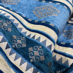 May include: Close-up of a folded fabric featuring a repeating pattern of blue, teal, and beige stripes. The design incorporates geometric shapes and textured details, creating a layered effect. The fabric appears soft and plush.