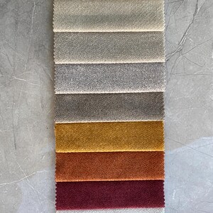 Op de afbeelding: Een verticale staal van stoffen in verschillende kleuren. De bovenste stalen zijn neutrale tinten, die overgaan in tinten goud, oranje en bordeaux. De stof heeft een getextureerde uitstraling, waarschijnlijk voor bekleding of woondecoratie.