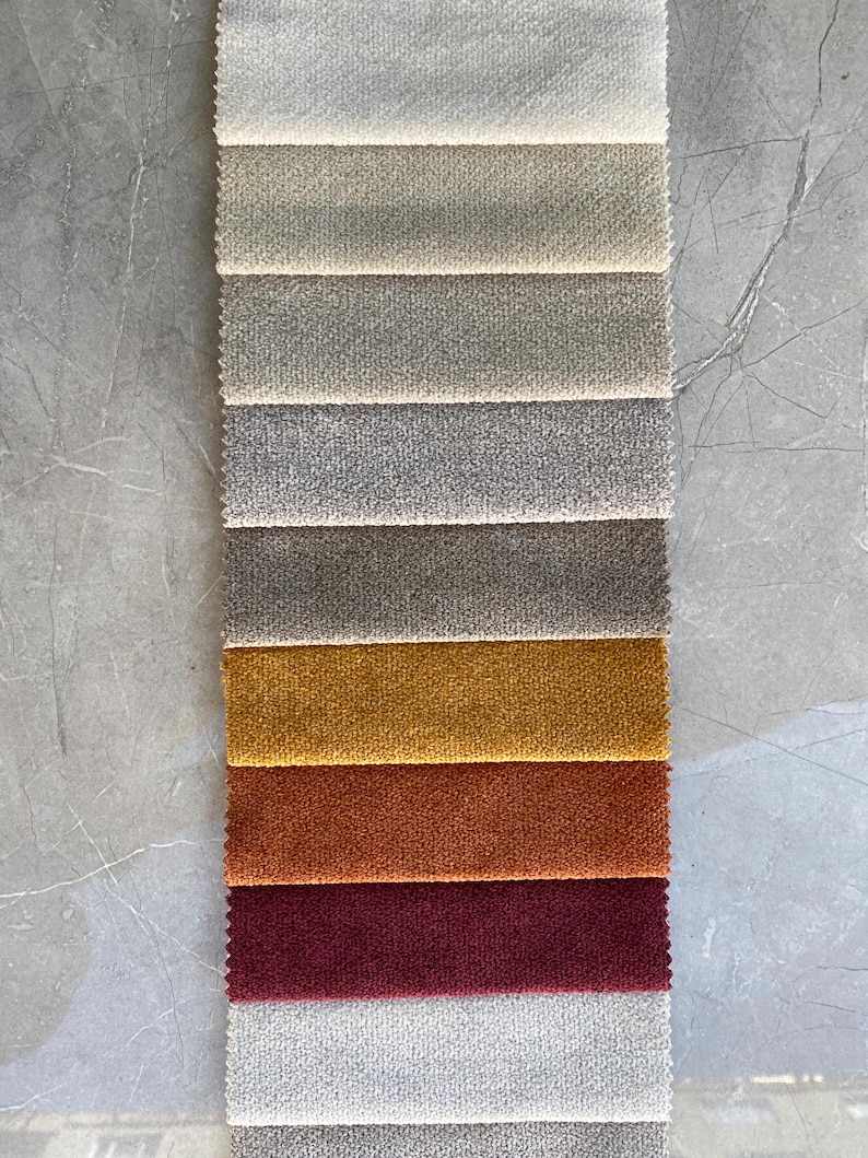 Op de afbeelding: Een verticale staal van stoffen in verschillende kleuren. De bovenste stalen zijn neutrale tinten, die overgaan in tinten goud, oranje en bordeaux. De stof heeft een getextureerde uitstraling, waarschijnlijk voor bekleding of woondecoratie.