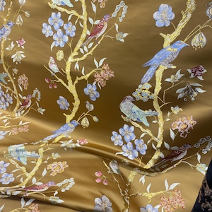 May include: Gold-colored fabric with a detailed pattern of birds, flowers, and branches. The birds are in shades of blue, red, and teal, while the flowers are light blue and pink. The branches are gold, creating a luxurious look.