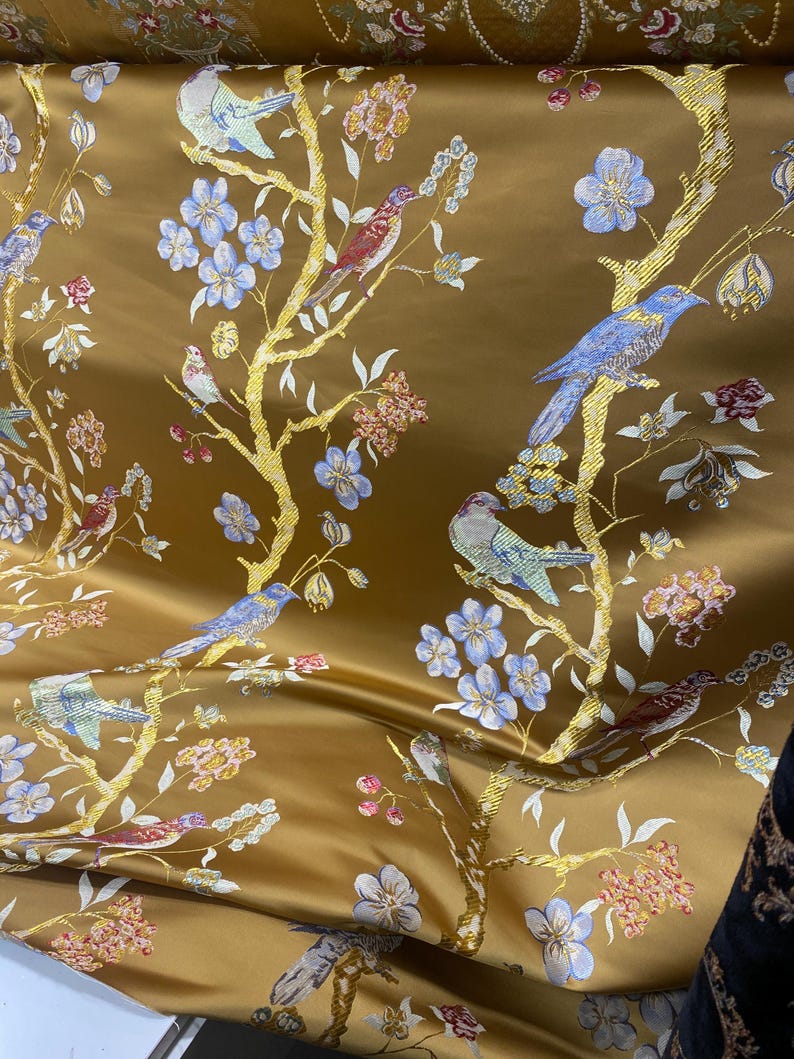 May include: Gold-colored fabric with a detailed pattern of birds, flowers, and branches. The birds are in shades of blue, red, and teal, while the flowers are light blue and pink. The branches are gold, creating a luxurious look.