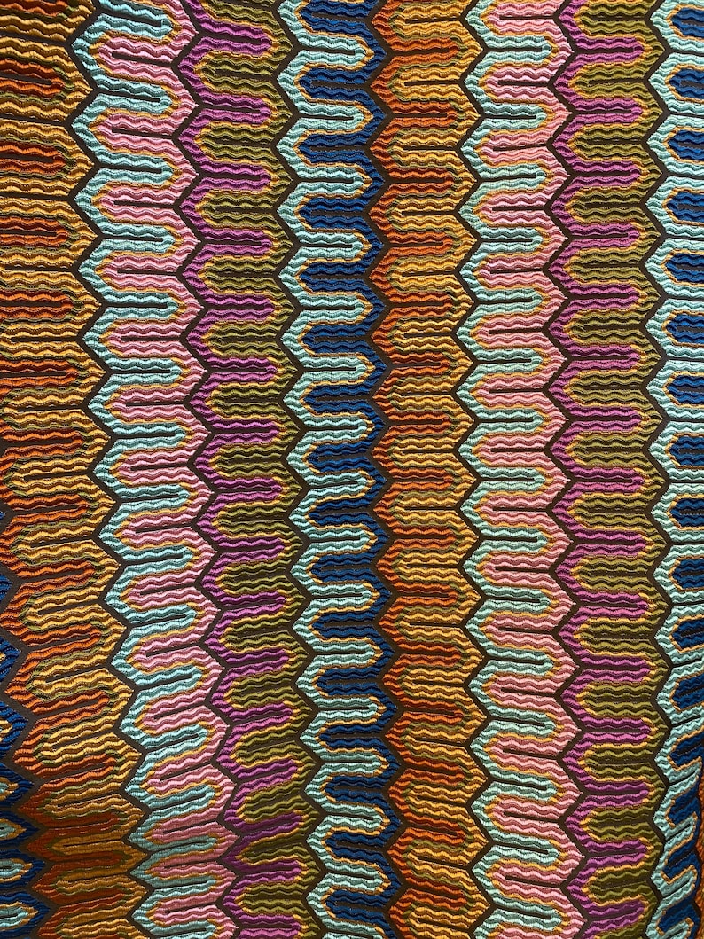 May include: Close-up of a colorful textile with a repeating geometric pattern. The design features a series of interconnected, wavy lines within hexagonal shapes. Colors include shades of blue, orange, pink, and purple, creating a vibrant and textured appearance.