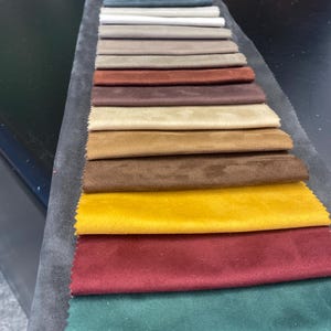 May include: A fabric swatch card featuring a variety of colors. The samples include shades of gray, blue, beige, brown, yellow, red, and green. The fabric appears to be a soft, velvety material, ideal for upholstery or textile projects.