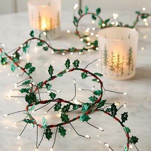 May include: A string of fairy lights with green holly leaves wrapped around the wire. The lights are strung around two clear glass candle holders with a white background.