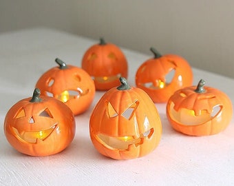 May include: Six orange pumpkin decorations with carved faces and green stalks. Each pumpkin has a light inside, creating a warm glow. These Halloween-themed decorations are arranged on a white surface.