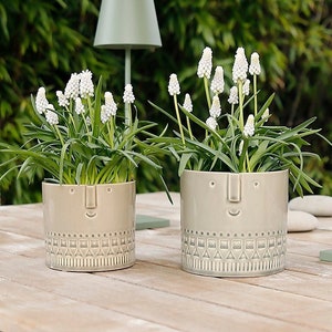 May include: Two green ceramic planters with a face design and geometric patterns. The planters are filled with white flowers and green plants. The planters are sitting on a wooden table with two green lamps in the background.