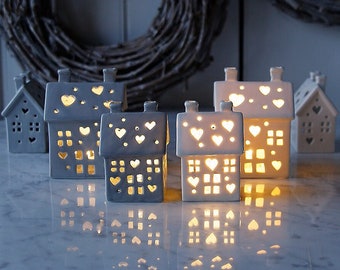 May include: Five white ceramic houses with cut-out hearts and windows. The houses are lit from within, casting a warm glow. The houses are arranged on a white marble surface.