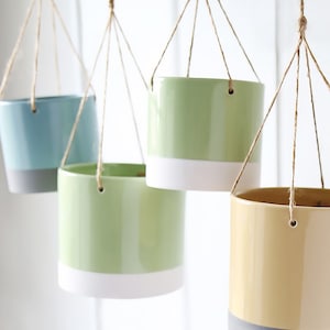 May include: Four hanging ceramic planters with a white rim. The planters are light blue, light green, light green, and yellow. The planters are hanging from twine.
