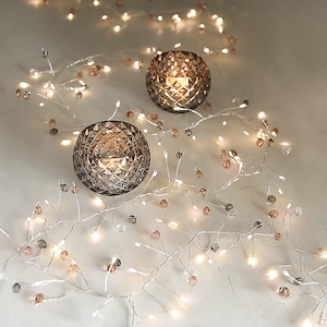 May include: A string of white fairy lights with clear and rose gold beads. The lights are wrapped around two clear glass candle holders.