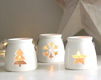 May include: Three white ceramic candle holders with cut-out designs. The candle holders feature a snowflake, a Christmas tree, and a star.