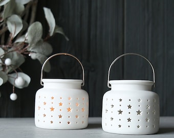 May include: Two white ceramic candle holders with star cutouts and metal handles. The candle holders are on a white surface.