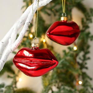 May include: Two red, glossy lip-shaped Christmas ornaments hanging from a white branch. The ornaments have a gold cap and are suspended by gold string. The background features green pine branches and soft, out-of-focus lights.