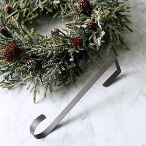 May include: A festive Christmas wreath made of green faux pine needles, decorated with brown pinecones, and a brown metal stocking holder. The wreath has white accents. The stocking holder is a simple, curved design.