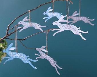 May include: Six wooden hare ornaments hanging from a branch. The hares are painted in shades of blue and gray with white floral designs. They are all leaping in mid-air.