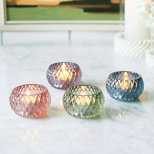 May include: Four glass votive candle holders in pink, purple, green, and blue. Each has a diamond-cut pattern and a lit candle. The holders are on a white marble surface, creating a soft, warm glow. Ideal for home decor.