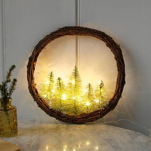May include: A circular, woven wreath featuring a ring of miniature, illuminated Christmas trees. The trees are lit with small, warm white lights. A small, yellow glass vase with a sprig of greenery is to the left of the wreath.