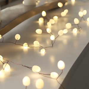 May include: A string of warm white fairy lights with round bulbs, draped over a wooden surface. The lights are strung on a thin copper wire.
