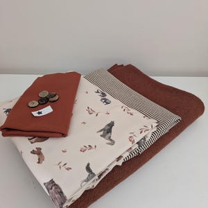 Könnte beinhalten: Ein Stapel gefalteter Stoffe, bestehend aus einem einfarbigen rostfarbenen Textil, einem cremefarbenen Stoff mit einem Muster aus kleinen Waldtieren und Blättern, einem braun-weiß gestreiften Stoff und einem dunkelbraunen Textil mit fünf runden Knöpfen.