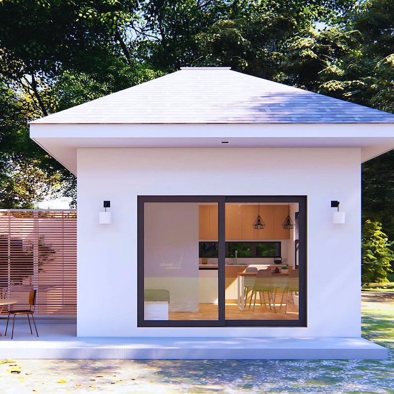 May include: A modern, white house with a gabled roof and large sliding glass doors. The interior is visible, showcasing a kitchen and dining area. Exterior features include a patio, wooden fence, and two wall-mounted lights. Lush green trees provide a natural backdrop.