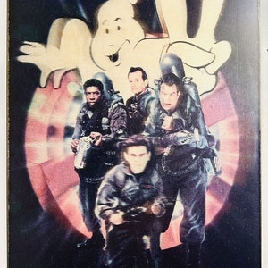 May include: A framed print featuring the Ghostbusters team and the iconic Slimer ghost. The image has a dark background with a pink and white circular design. The Ghostbusters are wearing their uniforms and holding proton packs.
