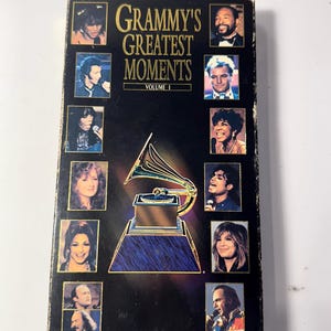 May include: A vintage VHS tape titled "Grammy's Greatest Moments Volume I." The black tape features a gold and blue graphic of a gramophone and portraits of various musical artists. The tape is rectangular and has a worn appearance.
