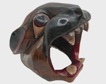 May include: A hand-carved wooden mask depicting a snarling jaguar head with an open mouth, sharp teeth, and prominent fangs. The mask features a glossy finish with black, brown, and deep red painted details, showcasing intricate facial markings.