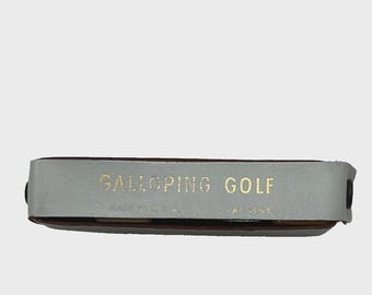 May include: A rectangular, light gray leather golf accessory with the words "GALLOPING GOLF" in gold lettering. The item is brown on the bottom and has a black button on the side. The text "MADE IN U.S.A." is also visible.