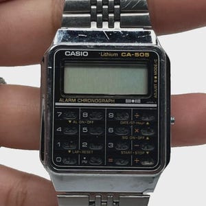 May include: Silver Casio CA-505 digital watch with a rectangular face and a stainless steel band. The watch features an alarm chronograph and various function buttons. The watch band is made of small, connected metal rectangles.