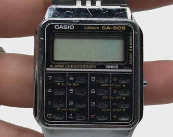 May include: Silver Casio CA-505 digital watch with a rectangular face and a stainless steel band. The watch features an alarm chronograph and various function buttons. The watch band is made of small, connected metal rectangles.