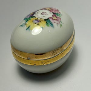 May include: A white porcelain egg-shaped trinket box with a gold trim and a floral design painted on the top.