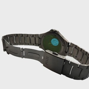 May include: A dark grey FUJI watch with a metal strap. The watch face is green with a blue sticker. The strap is open, showing the clasp and the FUJI logo.