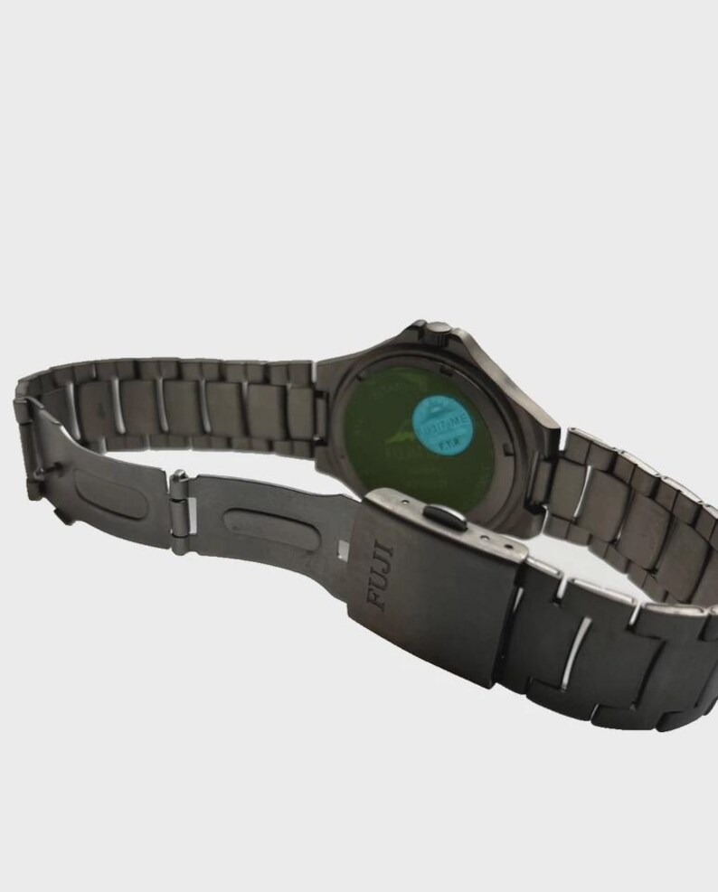 May include: A dark grey FUJI watch with a metal strap. The watch face is green with a blue sticker. The strap is open, showing the clasp and the FUJI logo.