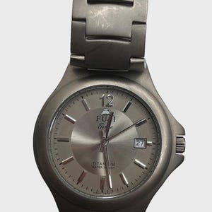 May include: A silver-toned FUJI Time wristwatch with a matching strap. The watch face has a date display at the 3 o'clock position and the words "TITANIUM WATER RESIST" are visible. The watch has a brushed metal finish.