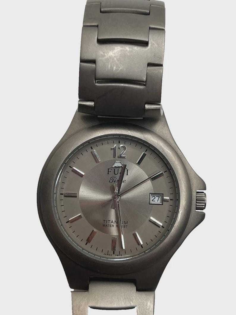 May include: A silver-toned FUJI Time wristwatch with a matching strap. The watch face has a date display at the 3 o'clock position and the words "TITANIUM WATER RESIST" are visible. The watch has a brushed metal finish.