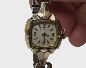 May include: A vintage Bulova wristwatch with a gold-toned case and band. The watch face is white with black numerals and hands. The band is a flexible, textured design. The watch is held in a hand.