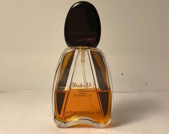 May include: A clear glass perfume bottle with a dark brown cap. The bottle has a unique shape with a curved design and contains an orange-colored liquid. The label reads "Balenciaga" and "Eau de Toilette".