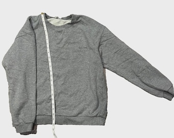May include: A gray sweatshirt with the word "CELINE" in black lettering. The sweatshirt has a crew neck and long sleeves. A measuring tape is placed on the sweatshirt. The sweatshirt is made of a soft material.